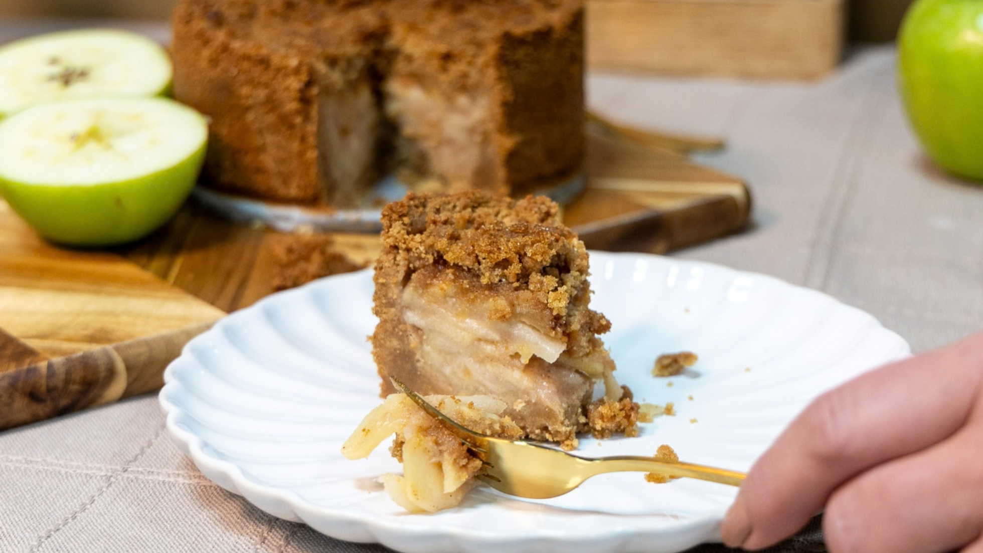 Slice of apple cake on a plate with a hand holding a fork, surrounded by apples and the rest of the cake.